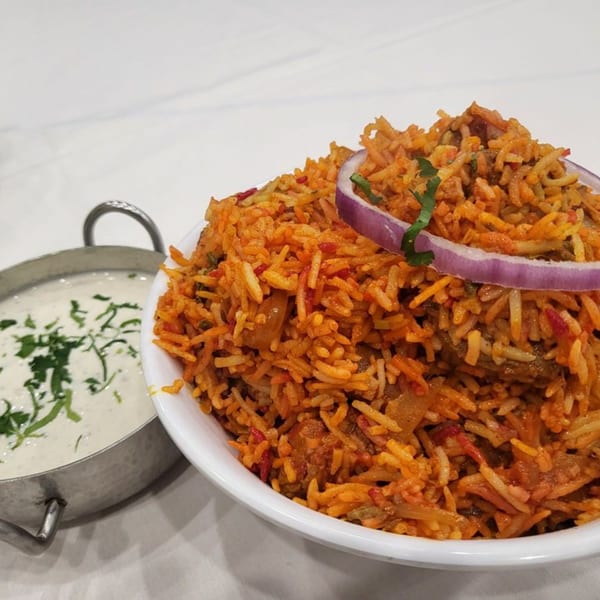 A bowl of orange-hued Indian biryani with red onion and cilantro, next to a metal bowl of raita.